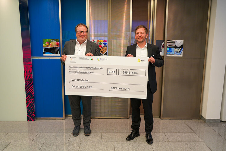 Minister Krischer und ein weiterer Mann halten einen übergroßen Förderscheck über 1.395.518,64 Euro in die Höhe