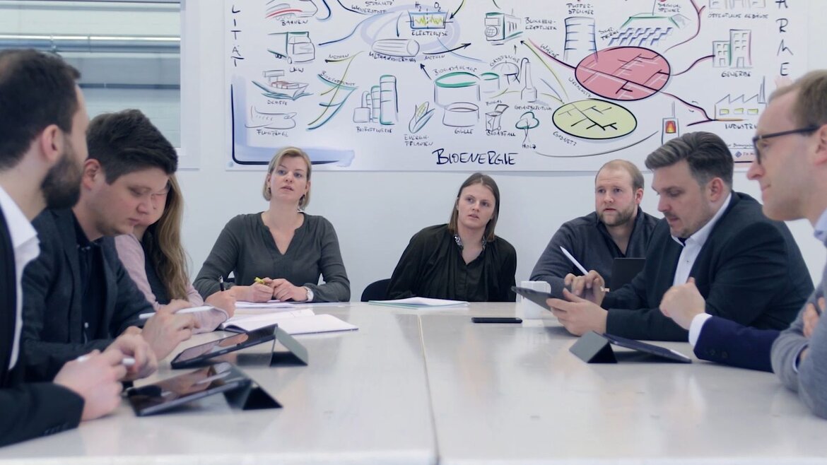 Mehrere Personen sitzen an einem Konferenztisch, im Hintergrund umfangreiche Skizzen zur Bioenergieversorgung