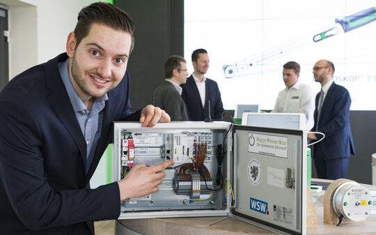 Ein junger Mann zeigt auf einen geöffneten Schaltkasten mit einem Smart Meter