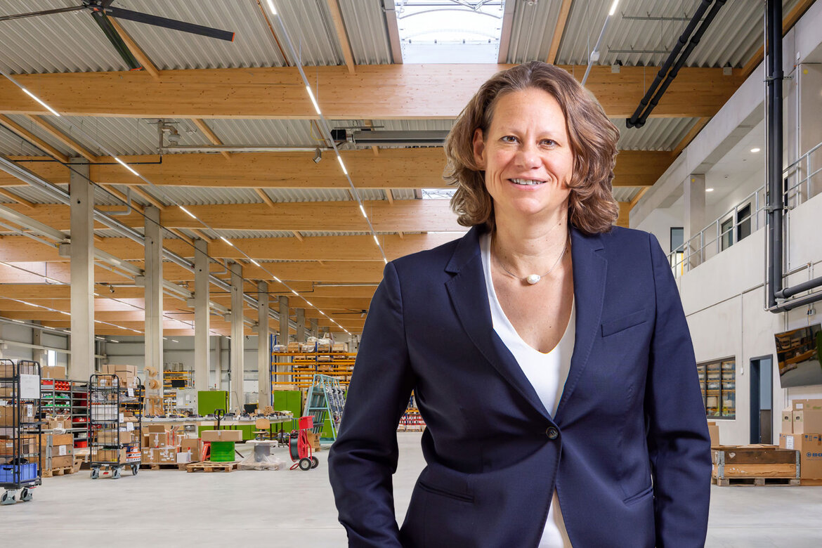 Frau Herzog in einem dunklen Blazer, im Hintergrund eine Industriehalle, tragende Elemente an der Decke sind aus Holz