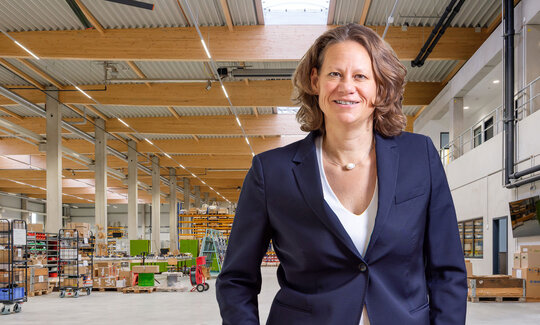 Frau Herzog in einem dunklen Blazer, im Hintergrund eine Industriehalle, tragende Elemente an der Decke sind aus Holz