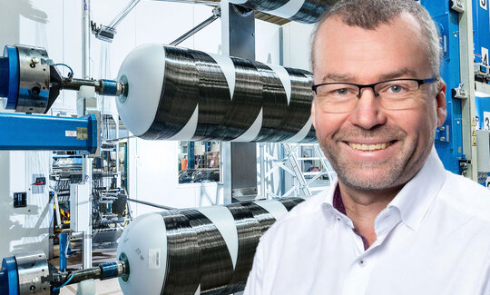 Ein Portrait von Wolfgang Wolter, im Hintergrund eine Maschine, die Kohlefasern zu Druckbehältern wickelt