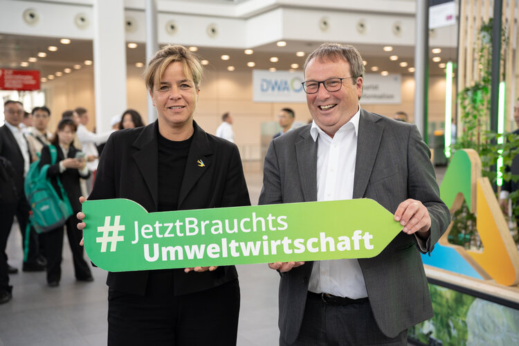 Ministerin Neubaur und Minister Krischer mit einem Pfeilobjekt und dem Hashtag #JetztBrauchtsUmweltwirtschaft