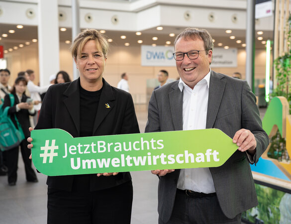 Ministerin Neubaur und Minister Krischer mit einem Pfeilobjekt und dem Hashtag #JetztBrauchtsUmweltwirtschaft
