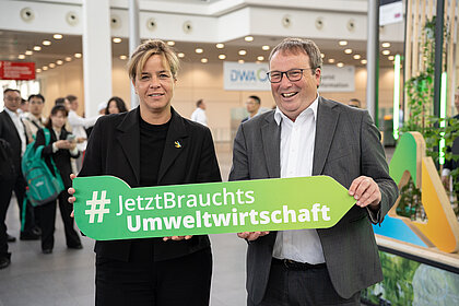 Ministerin Neubaur und Minister Krischer mit einem Pfeilobjekt und dem Hashtag #JetztBrauchtsUmweltwirtschaft