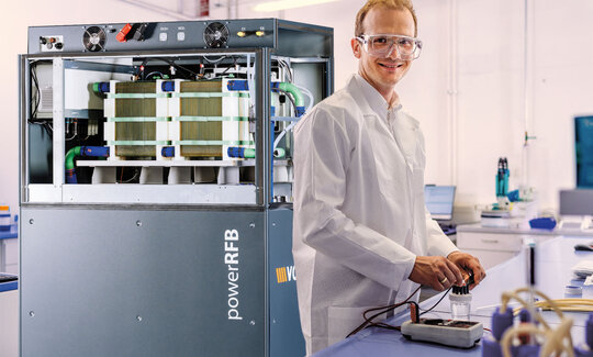 Ein Mann im Laborkittel steht an einem Labortisch, im Hintergrund steht eine große Redoxflow-Batterie