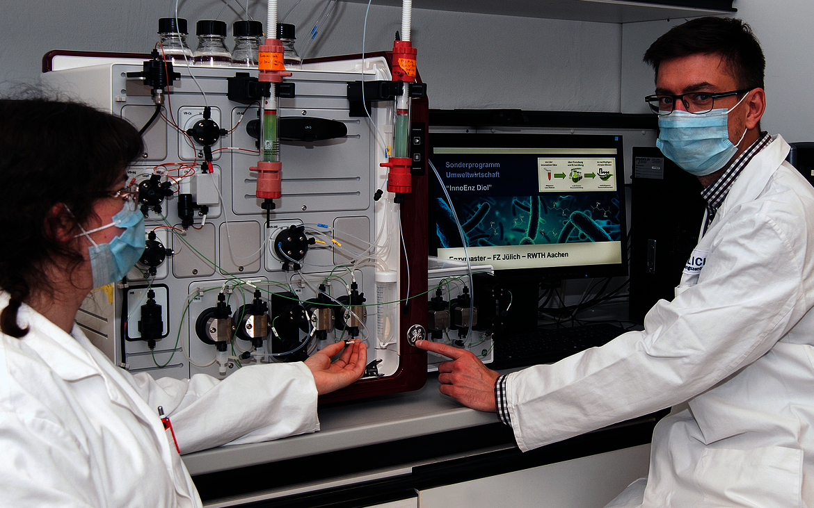 Ein Mann und eine Frau in Laborkleidung sitzen vor einer technischen Apparatur, im Hintergrund steht auf einem Schild "InnoEnz Diol", Sonderprogramm Umweltwirtschaft