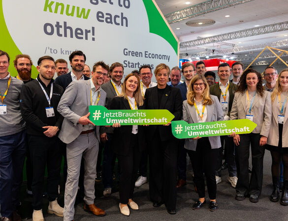 Gruppenbild mit Ministerin Neubaur vor einem Messestand, mehrere Personen halten das Pfeilsymbol #JetztBrauchtsUmweltwirtschaft
