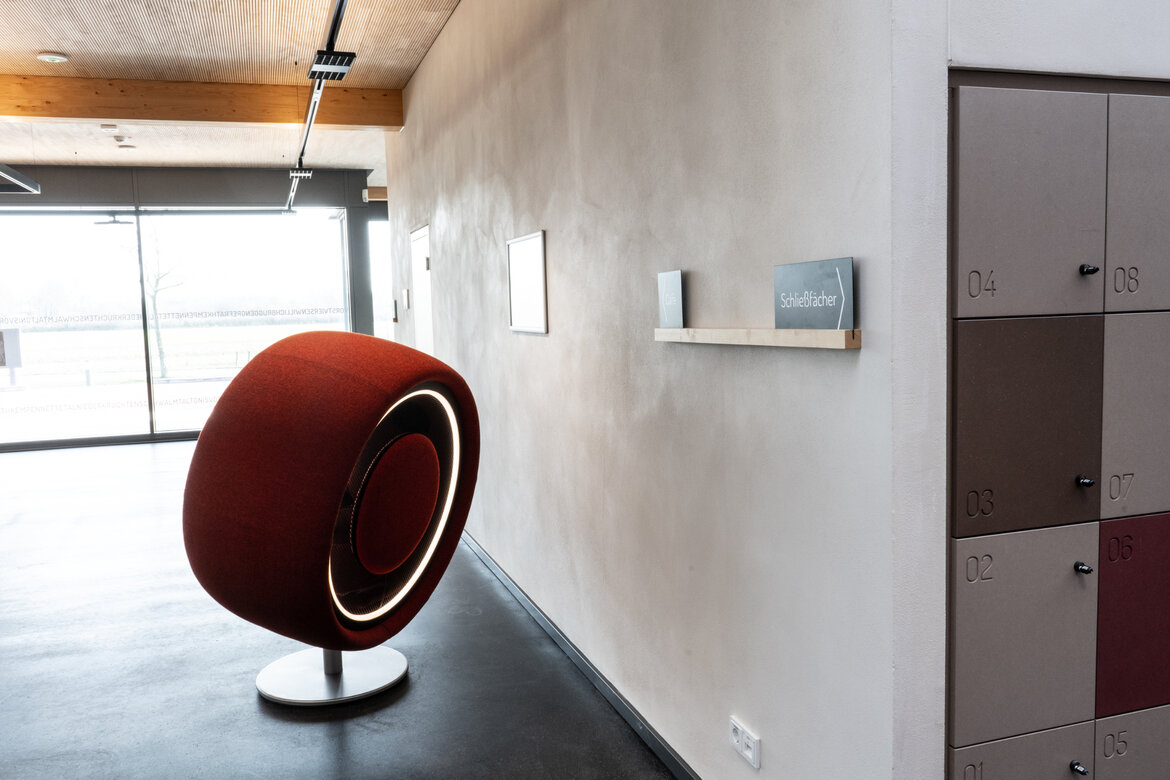 Ein moderner, heller Publikumsbereich, ein Designersessel steht an einer mit Lehm verputzten Wand