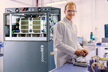 Ein Mann im Laborkittel steht an einem Labortisch, im Hintergrund steht eine große Redoxflow-Batterie